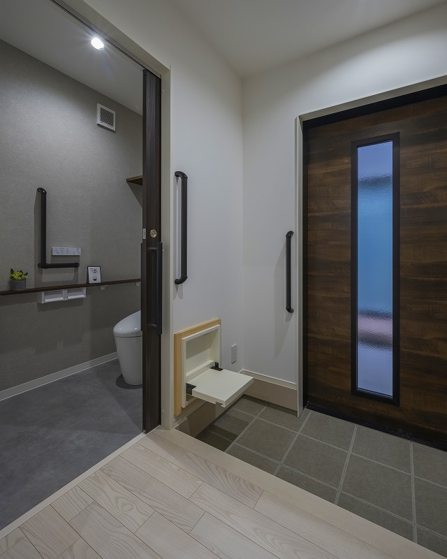 落ち着いた色味の玄関扉が印象的な玄関空間。靴の脱ぎ履きをサポートする折り畳み式ベンチに加え、立ち座りを補助する手すりを設置しました。玄関横にトイレを配置することで、帰宅後や外出前もスムーズな動線を確保しています。