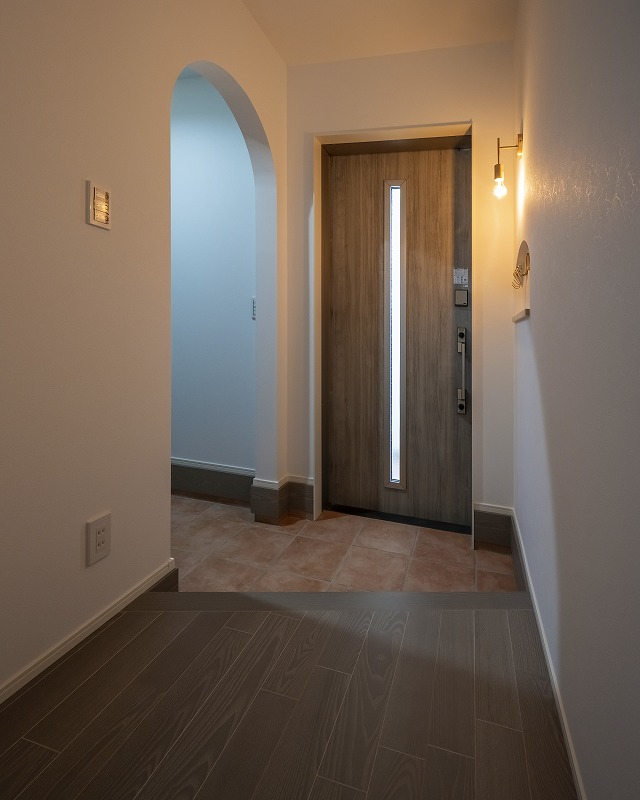 アーチ垂れ壁がやさしい雰囲気を演出する玄関。木目調のドアやタイルの床、シンプルな照明が落ち着いた空間をつくります。玄関横にはシューズクロークへとつながる動線もあり、毎日の使い勝手を両立しました。